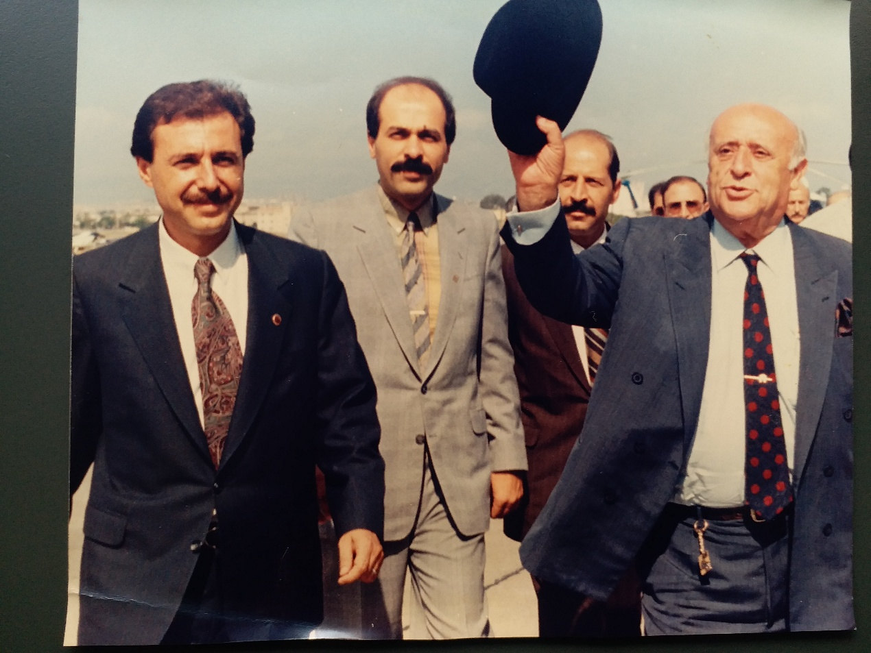 Süleyman Demirel, havalimanı apronunda şapkasını kaldırarak selam veriyor; yanında takım elbiseli karşılama heyeti yürüyor.